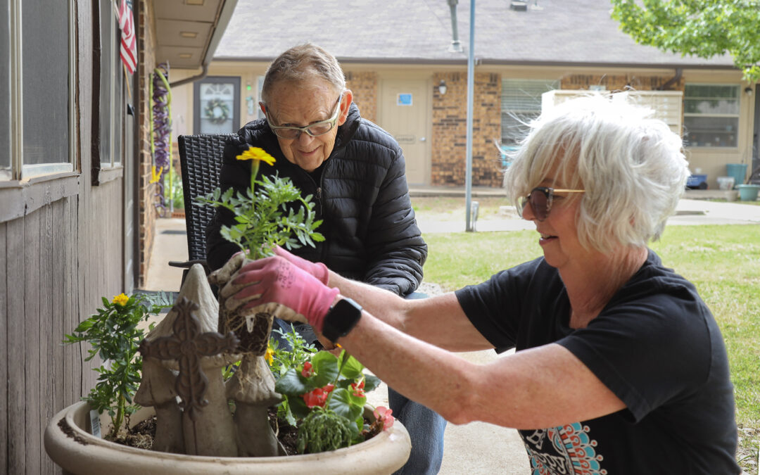 A woman plants flowers in a pot as a man watches.