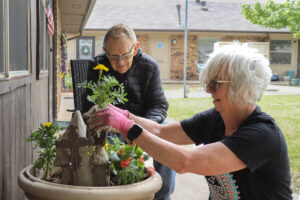 A woman plants flowers in a pot as a man watches.